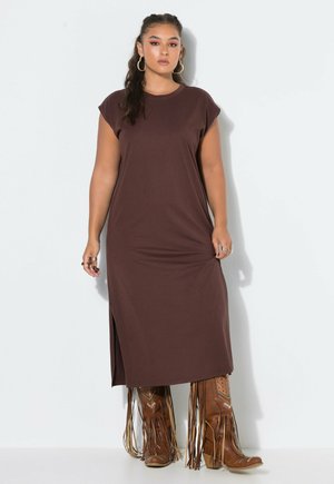 Jeune femme aux longs cheveux foncés porte une robe midi marron sans manches et de hautes bottes en cuir marron à franges, debout devant un fond blanc uni.