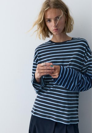 Jeune femme aux cheveux blonds courts portant un chemisier rayé bleu marine et bleu clair à manches longues, les mains jointes près de la poitrine.