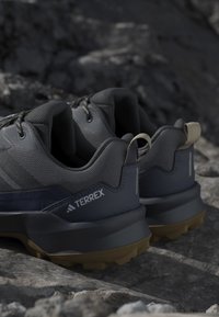 Coppia di scarpe da trail Adidas Terrex grigio scuro e nero con suole marroni poggiate su terreno roccioso.