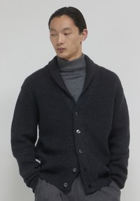 Uomo che indossa un cardigan a coste grigio scuro con bottoni sopra un dolcevita grigio, con le mani nelle tasche, che guarda di lato contro uno sfondo semplice.