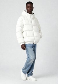 Giacca imbottita bianca con cappuccio, caratterizzata da un design trapuntato e polsini elastici, abbinata a jeans azzurri e scarpe da ginnastica bianche.