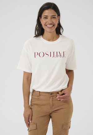 Femme souriante avec des cheveux foncés portant un t-shirt blanc avec l'inscription "Vibes POSITIVES" et un pantalon cargo beige devant un fond uni.