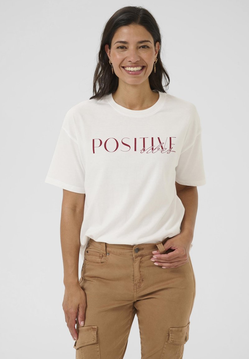 Usmívající se žena s tmavými vlasy, oblečená v bílém tričku s nápisem "POSITIVE vibes" a béžových cargo kalhotách, na jednobarevném pozadí.