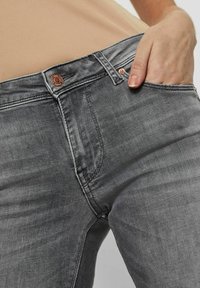 Los jeans de mezclilla gris presentan un corte ajustado, un diseño de cinco bolsillos y un botón de metal marrón. La tela muestra una ligera textura con áreas desgastadas.