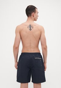 Marine blå svømmebukser laget av lett materiale, med elastisk livstropp, baklomme med dekorativ trim, og en avslappet passform.