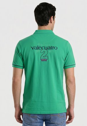 Hombre con camisa polo verde con texto "valecuatro 2" en la espalda y vaqueros azules, de pie frente a un fondo blanco liso.