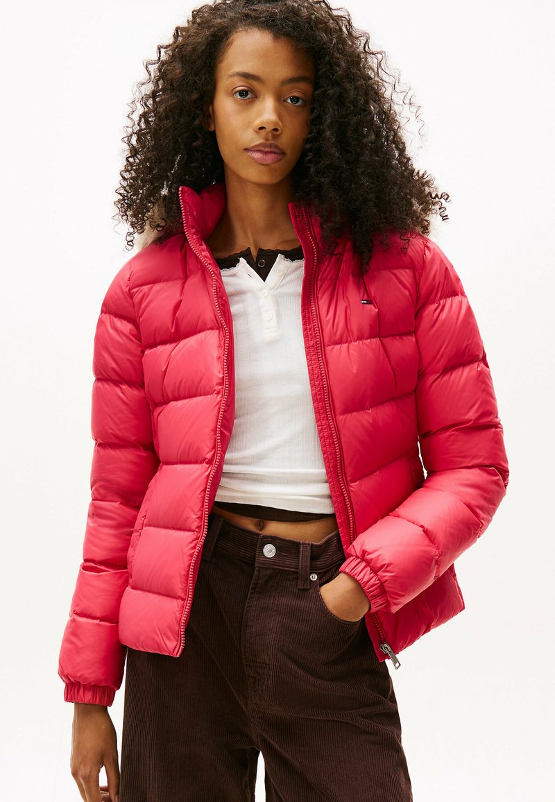 Czerwona kurtka puchowa o pikowanym designie i wysokim kołnierzu. Noszona na białej bluzce i brązowych spodniach sztruksowych, podkreślająca dopasowaną sylwetkę.