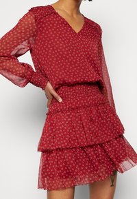 Robe à motif rouge avec des manches longues transparentes, décolleté en V, taille smockée et jupe à volants superposés dotée de petits motifs floraux.