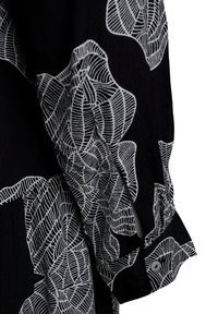 Schwarzes Stoffhemd mit weißem Blumendesign. Lange Ärmel mit Knopfmanschetten-Detail. Strukturierte Material verstärkt den visuellen Kontrast.