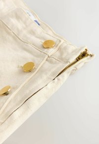 Beige denim stof, met een gouden rits en drie ronde gouden knoopsluitingen langs de bovenrand, met zichtbare stikseldetails.