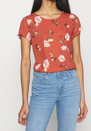 Blusa color ruggine con maniche corte arrotolate, caratterizzata da un motivo floreale in rosa e crema, abbinata a jeans azzurri chiari.