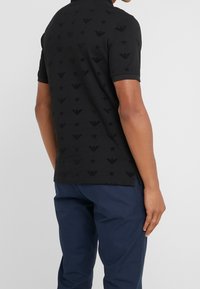 Svart polo-tröja med en strukturerad mönstring av logotyper och stjärnor, med krage och korta ärmar, i kombination med marinblå byxor.