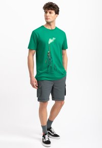 Groene katoenen t-shirt met een bedrukt ontwerp op de voorkant, gecombineerd met grijze cargoshorts. Het model draagt zwarte sneakers en grijze sokken.