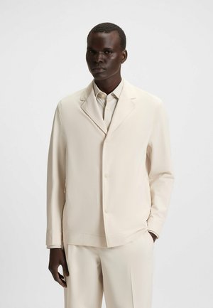 Beige lightweight jacket with a single button closure, notch lapel, and straight cut. Paired with matching beige pants. Smooth texture.