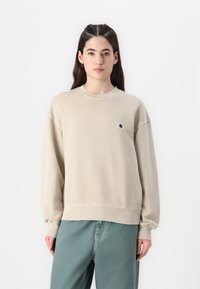 Felpa beige con colletto rotondo e polsini a costine. Presenta un piccolo patch logo sul petto. Abbinata a pantaloni verdi larghi.