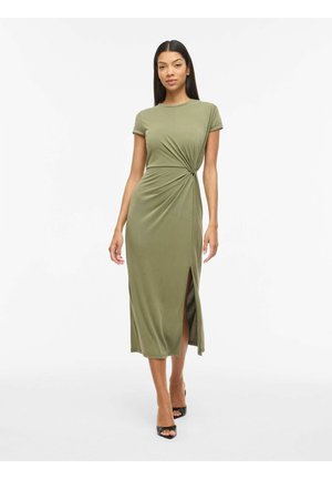 Kobieta ubrana w oliwkową sukienkę midi z krótkim rękawem, rozcięciem po boku i marszczeniem w talii, dobraną do czarnych sandałów na otwartym palcu.