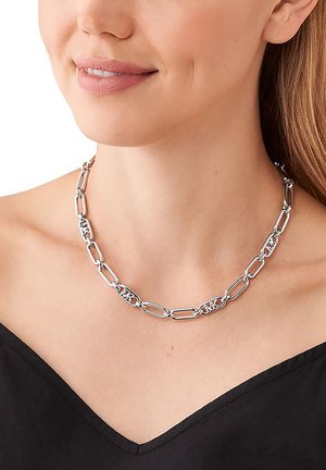 Silverkedja med avlånga, sammanlänkade länkar med en texturerad design. Bärs på en bar hals ovanför en svart topp.