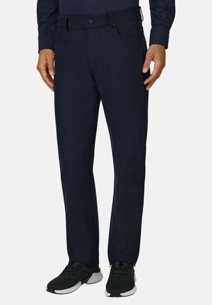 Homme portant un pantalon droit bleu marine foncé avec des chaussures de sport noires et blanches, debout devant un fond uni.