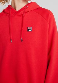 Primer plano de una persona que lleva una sudadera roja brillante con cordones y un pequeño logo de FILA en el pecho.