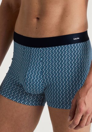 Blaue gemusterte Boxershorts mit einem Zickzackdesign, ausgestattet mit einem dunklen elastischen Bund, der mit "CALIDA" beschriftet ist. Hergestellt aus einem glatten, dehnbaren Material.
