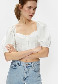 Weißes Oberteil mit Puffärmeln und strukturiertem Oberteil, ausgestattet mit texturiertem Stoff und Knopfverschluss vorne. Kombiniert mit hellblauen High-Waist-Jeans.