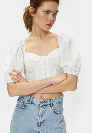 Weißes Oberteil mit Puffärmeln und strukturiertem Oberteil, ausgestattet mit texturiertem Stoff und Knopfverschluss vorne. Kombiniert mit hellblauen High-Waist-Jeans.