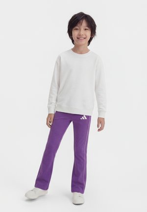 Usmievajúce sa dieťa stojace oblečené v bielej tričku s dlhým rukávom, fialových teplákoch Adidas a bielych teniskách na jednofarebnom pozadí.
