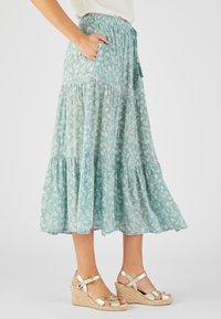 Jupe maxi bleu sarcelle clair avec imprimé floral blanc, design à volants, taille élastique et poches latérales. Associée à des sandales compensées dorées.
