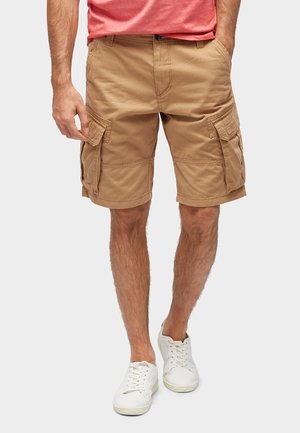 Ein Mann trägt beige Cargoshorts und weiße Turnschuhe, steht mit den Händen teilweise in den Taschen vor einem schlichten Hintergrund.