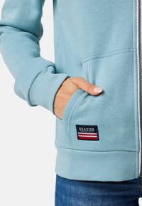 Lichtblauwe rits hoodie met een zachte textuur, voorzien van een voorzak en een 'SEASIDE' logo patch met rode en witte strepen.