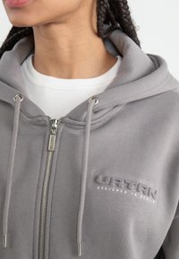 Grå hettejakke med glidelås og snøring, mykt stoff, sølvfarget hardware, preget logo, og en hvit ribbestrikket skjorte under.