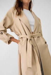 Trench-coat beige avec une taille ceinturée, de grands revers et des détails de poignets boutonnés. Texture lisse, coupe longueur genou.