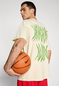 Ung mand, der holder en orange basketball under armen, iført en cremefarvet skjorte med grøn grafisk tekst på ryggen og røde shorts.