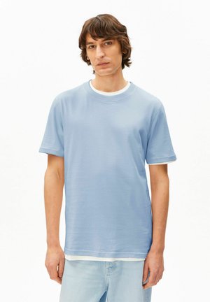 Junger Mann mit gewelltem braunem Haar, der ein hellblaues kurzärmeliges T-Shirt über einem weißen Hemd und hellblaue Jeans trägt, stehend vor einem weißen Hintergrund.