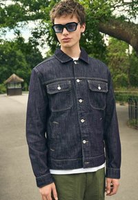 Mørk indigo denimjakke med lodrette striber, to brystlommer og hvide knapper. Båret over en hvid skjorte og parret med grønne bukser.