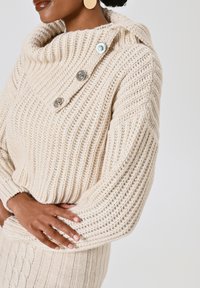 Maglione beige lavorato a maglia con colletto a cappuccio, caratterizzato da tre grandi bottoni argentati e una texture a coste. Maniche ampie e fondo aderente.
