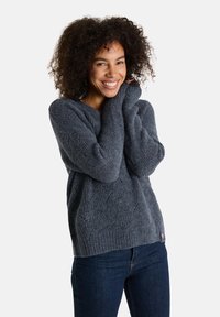 Grauer Strickpullover mit strukturiertem Muster, gerippten Bündchen und lockerer Passform. Getragen mit dunkelblauen Jeans und mit einem kleinen Logo-Detail.