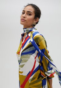 Camicia a fantasia colorata con vivaci tonalità di giallo, blu, rosso e verde. Presenta un colletto e maniche lunghe con dettaglio a fiocco.