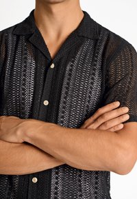 Camisa negra de manga corta con un patrón abierto de punto, que presenta rayas verticales y botones claros, mostrando un diseño texturizado.