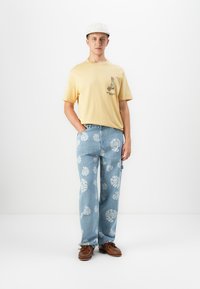 Ung mand iført en lys gul T-shirt med kyllinge-tryk, lyseblå jeans med hvide spiral mønstre, hvid kasket og brune sko.