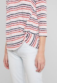 Gestreiftes Oberteil in Rot und Marineblau mit einem geknoteten Seitendetail. Aus leichtem Stoff gefertigt. Mit Dreiviertelärmeln und einem geschwungenen Saum, kombiniert mit hellen Jeans.