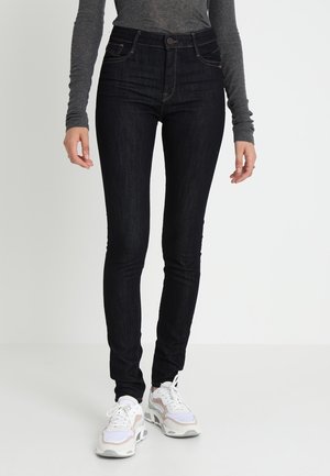 Jeans de mezclilla negros de tiro alto con una silueta ajustada, que presentan costuras en contraste y un diseño de cinco bolsillos, combinados con zapatillas en tonos neutros.