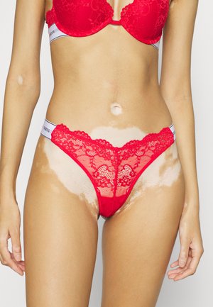 Rode kanten string met ingewikkelde bloemmotieven, elastische band met branding en een semi-doorzichtig ontwerp, getoond tegen een neutrale achtergrond.