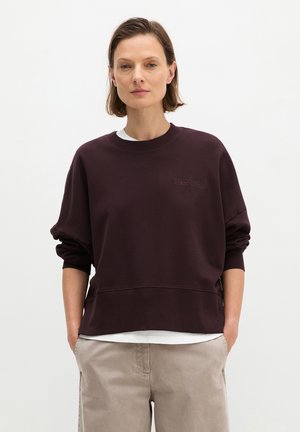 Vrouw met kort bruin haar, gekleed in een donkerbruine trui over een wit shirt en beige broek, met handen in de zakken, staand tegen een effen achtergrond.
