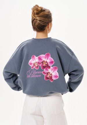 Kobieta o jasnobrązowych włosach związanych w kok, ubrana w szarą bluzę z różowymi kwiatami orchidei oraz napisem „Bloom in silence” na plecach.