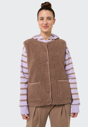 Frau mit Haaren zu einem Dutt gebunden, trägt eine braune flauschige Weste über einem helllila und braun gestreiften Hoodie und braune Hosen, lächelt leicht.