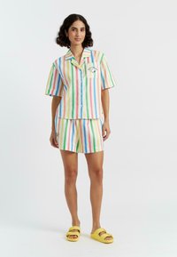 Completo pigiama a maniche corte a righe in colori pastello (rosa, blu, verde, giallo) con taschino sul petto e dettagli ricamati; abbinato a sandali gialli.