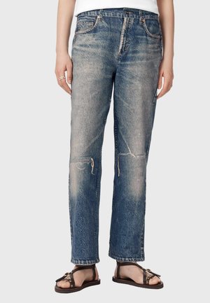 Person iført falmede, slidte blå jeans og brune sandaler med åben tå, stående mod en ensfarvet baggrund.