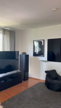 Televisão preta num móvel baixo, sistema de som preto elegante e uma cadeira de feijão escura sobre um tapete cinza texturizado. A arte na parede apresenta uma moldura reflexiva.