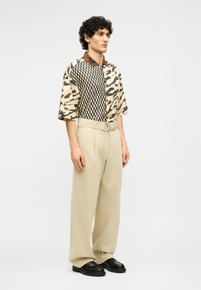 Άνδρας με σγουρά μαλλιά, που φορά κοντομάνικο πουκάμισο με ανάμεικτα σχέδια ζώων και σκακιέρας, μπεζ παντελόνια φαρδιάς γραμμής και μαύρα παπούτσια.